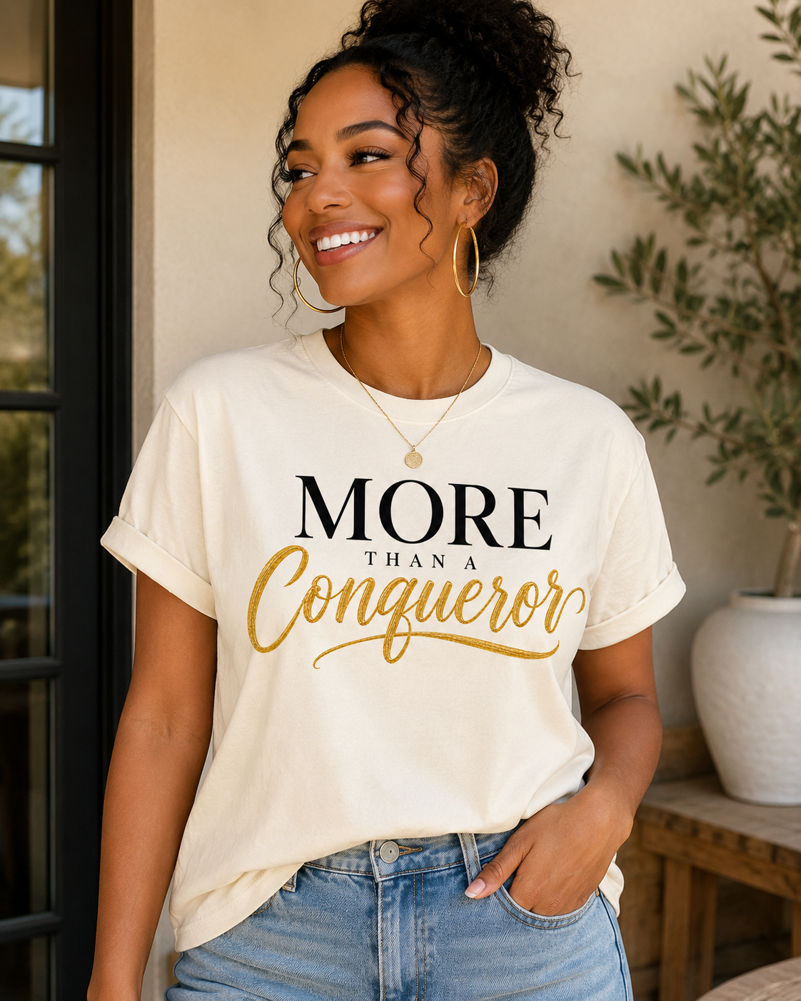 More Than A Conqueror Christian t-shirt in soft natural with bold black and gold script typography.